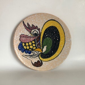 Puede incluir: Un plato de cerámica blanco con un diseño de gallo colorido. El gallo está pintado en tonos de amarillo, rojo, azul, verde y negro. El plato tiene un esmalte moteado.