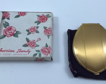 May include: A gold compact mirror with a rose design on the front. The mirror is in a purple velvet pouch. The compact is in its original box, which has a white background with pink roses and the text "American Beauty by Elgin American".