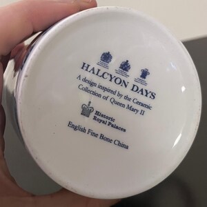 Puede incluir: Base de una taza de cer&aacute;mica blanca con texto y logotipos azules. El texto dice "HALCYON DAYS" y "A design inspired by the Ceramic Collection of Queen Mary II." Tambi&eacute;n incluye "Historic Royal Palaces" y "English Fine Bone China."