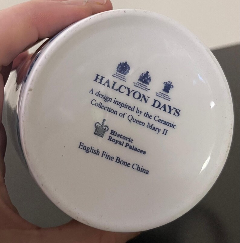 Puede incluir: Base de una taza de cer&aacute;mica blanca con texto y logotipos azules. El texto dice "HALCYON DAYS" y "A design inspired by the Ceramic Collection of Queen Mary II." Tambi&eacute;n incluye "Historic Royal Palaces" y "English Fine Bone China."