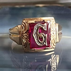 Peut inclure: Une bague dorée avec une pierre centrale rectangulaire rouge rubis. La pierre présente un « G » blanc stylisé. La bague a des détails ornés sur les côtés et une finition polie, reflétant la lumière.