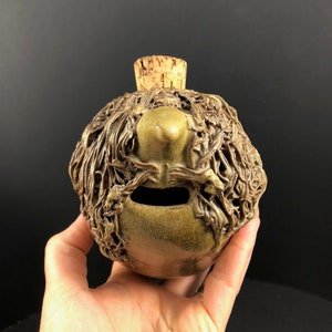 May include: A ceramic bottle with a cork stopper, shaped like a face with a long nose and a mouth. The bottle is covered in a brown glaze with a textured surface.