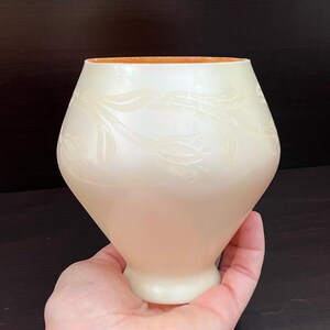 May include: Cream-colored vase with a tulip design. The vase has a wide, rounded body and a narrow base. The interior rim is a warm orange color. The tulip design is etched into the surface of the vase. The vase is held in a hand.