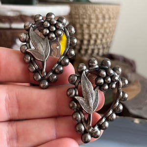 May include: A pair of silver tone clip-on earrings with a heart-shaped design. The earrings feature a cluster of small beads around the edge and a leaf design in the center.