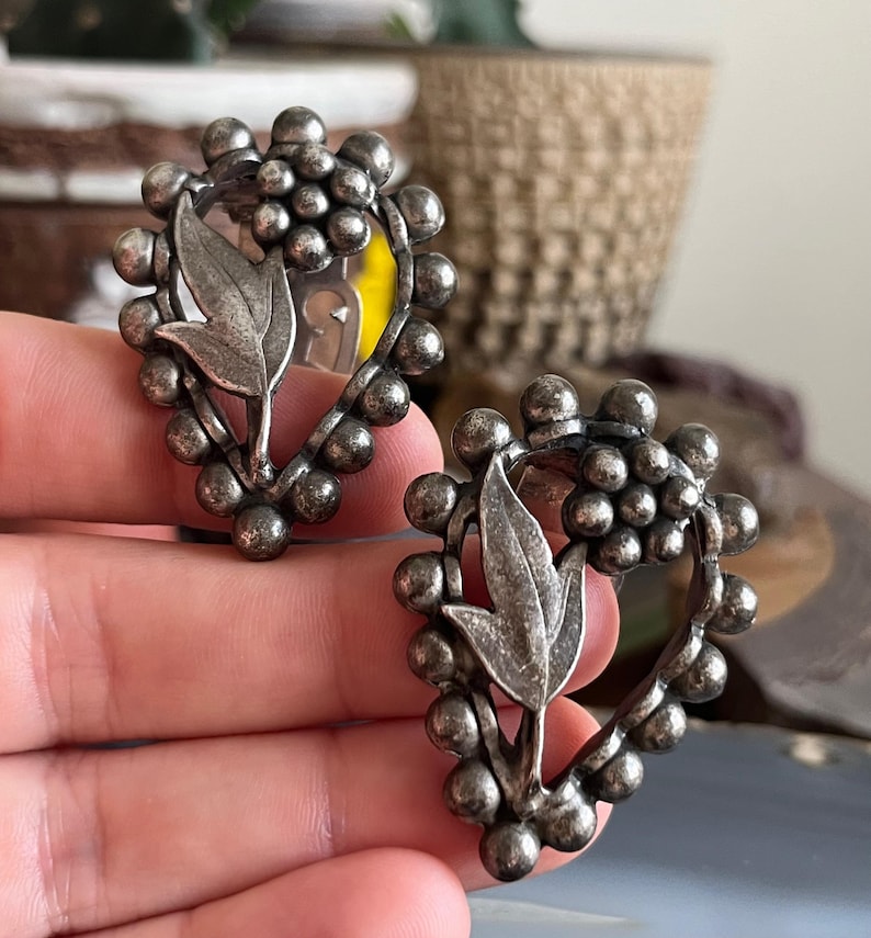May include: A pair of silver tone clip-on earrings with a heart-shaped design. The earrings feature a cluster of small beads around the edge and a leaf design in the center.
