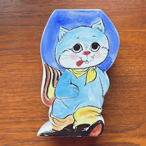 May include: A ceramic cat figurine with a blue body, a yellow scarf, and black shoes. The cat has a red tongue sticking out and is wearing a blue hat. The figurine is hand-painted with a cartoonish style.