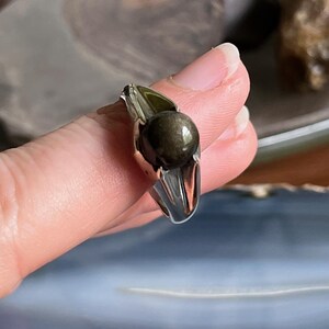 May include: A silver ring with a dark, round, iridescent stone. The ring's band is a polished silver, and the stone has a metallic sheen. The ring is held up, showcasing its design and the stone's color.