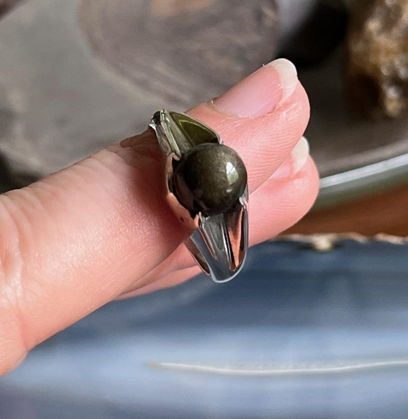 May include: A silver ring with a dark, round, iridescent stone. The ring's band is a polished silver, and the stone has a metallic sheen. The ring is held up, showcasing its design and the stone's color.