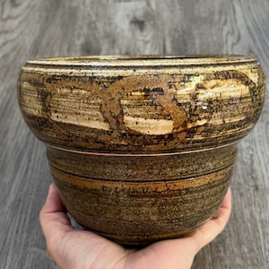 May include: A handmade ceramic planter with a wide, rounded top and a slightly narrower base. The planter has a textured surface with horizontal bands of brown, tan, and black, creating a rustic aesthetic. The planter is held by a hand.