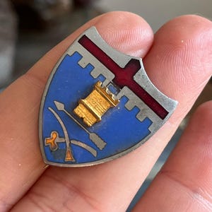 Pode incluir: Um broche de escudo prateado com fundo azul, uma cruz vermelha e uma torre dourada. O broche tem um fecho de broche prateado.