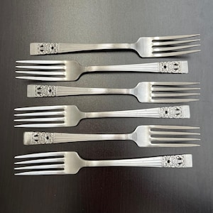 May include: Six silver forks with a decorative floral design on the handle. The forks are arranged on a dark surface, showcasing their tines and the detailed pattern on the handle.