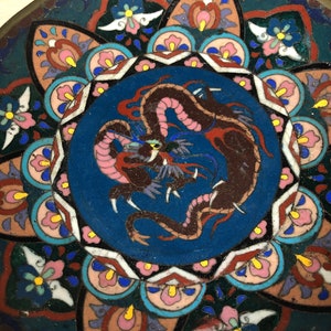 May include: A round, blue and white ceramic plate with a colorful dragon design in the center. The dragon is brown, pink, and red with a yellow eye. The plate is decorated with a floral pattern around the edge.