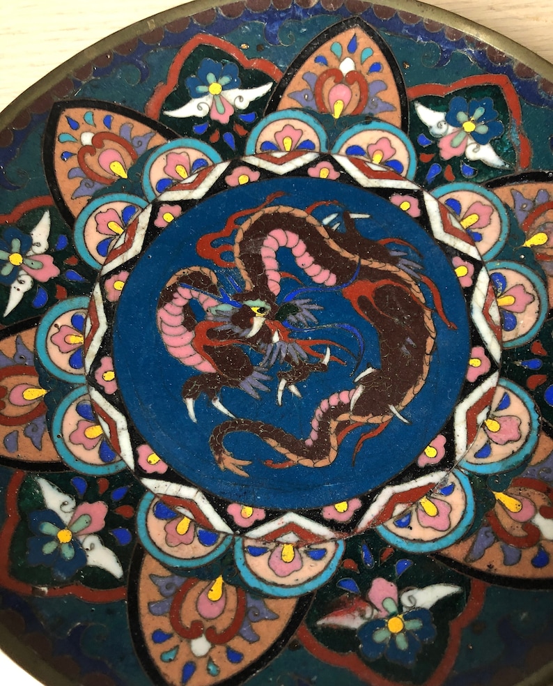 May include: A round, blue and white ceramic plate with a colorful dragon design in the center. The dragon is brown, pink, and red with a yellow eye. The plate is decorated with a floral pattern around the edge.