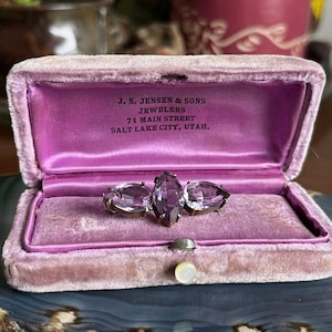 May include: A vintage amethyst brooch with three faceted stones, displayed in an open, pink velvet jewelry box. The box interior is lined with pink satin and features the text "J. S. Jensen & Sons Jewelers, 71 Main Street, Salt Lake City, Utah."