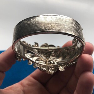 K&ouml;nnte beinhalten: Ein silbernes Armband mit einem detaillierten Blumendesign. Das Armband ist offen und hat eine geb&uuml;rstete Oberfl&auml;che.