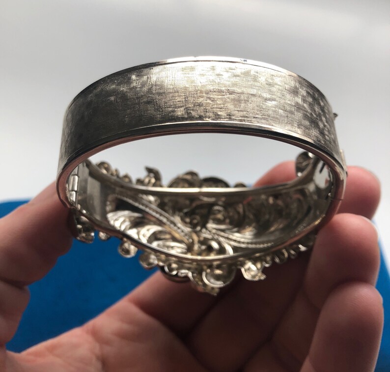 K&ouml;nnte beinhalten: Ein silbernes Armband mit einem detaillierten Blumendesign. Das Armband ist offen und hat eine geb&uuml;rstete Oberfl&auml;che.