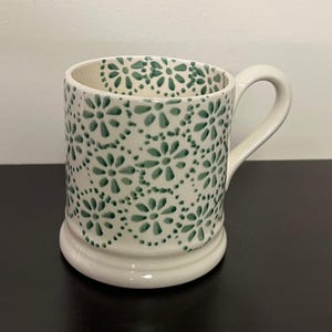 May include: White ceramic mug with a green floral pattern. The mug has a rounded base, a curved handle, and a detailed design of green flowers and dots. The interior of the mug is white.