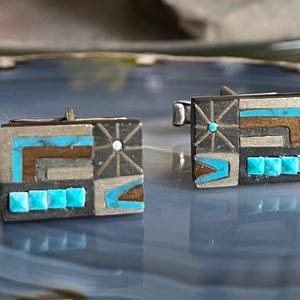 Puede incluir: Un par de gemelos cuadrados con un diseño geométrico. Los gemelos presentan una base de metal plateado con incrustaciones turquesas, marrones y plateadas. El diseño incluye cuadrados, rectángulos y un patrón de estrella.
