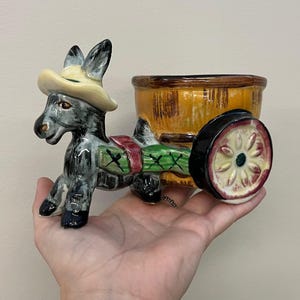 May include: A ceramic donkey figurine pulling a cart. The donkey is gray and black, wearing a cream-colored hat. The cart is yellow and brown with a black wheel. The donkey has a green and red saddle.