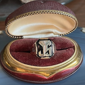 May include: A gold ring with a black onyx face, a diamond, and stylized initials. The ring is presented in an open, maroon velvet-lined jewellery box with a gold trim. The box is open, displaying the ring.