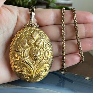 May include: A gold-colored oval pendant necklace with intricate floral and leaf engravings. The pendant hangs from a matching gold chain. The design features detailed patterns and textures, creating a vintage aesthetic.