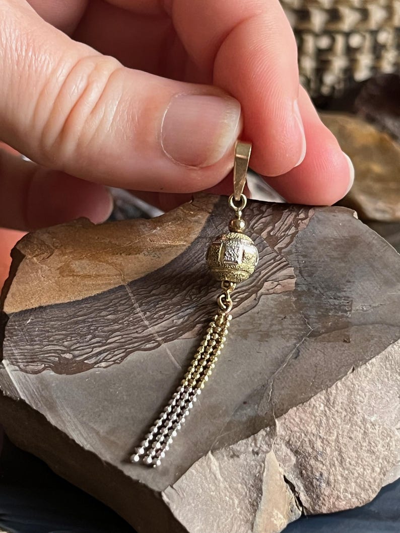 Puede incluir: Un colgante de oro y plata con una bola redonda texturizada y un borla de cadena.