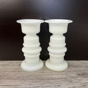 Op de afbeelding: Twee witte glazen kandelaars. Elk heeft een brede, ondiepe bovenkant, een cilindrisch lichaam met gestapelde, afgeronde secties en een cirkelvormige basis. De kandelaars worden gepresenteerd op een houten oppervlak met een donkere achtergrond.