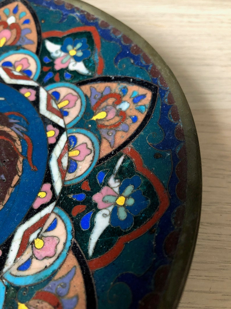 May include: A round, decorative plate with a blue, green, pink, and yellow floral pattern. The plate is made of metal and has a slightly raised rim.