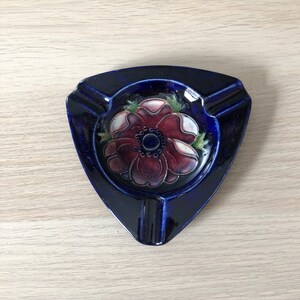 May include: A blue ceramic ashtray with a floral design. The flower is red, white, and green. The ashtray is triangular in shape.