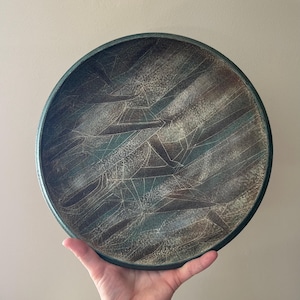 May include: A round ceramic plate with a dark teal rim and a textured surface. The plate features a pattern of brown and teal diagonal lines with white crackle lines throughout. The plate is held by a hand.