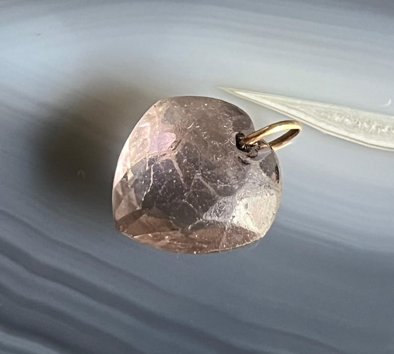 May include: A faceted, heart-shaped pendant in a light brown hue. The pendant has a gold-colored bail for attaching to a chain. The surface of the heart has multiple facets, giving it a textured appearance.