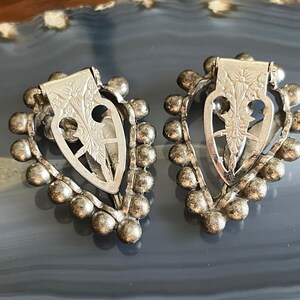 May include: A pair of silver heart-shaped earrings with a beaded border and a floral design in the center.