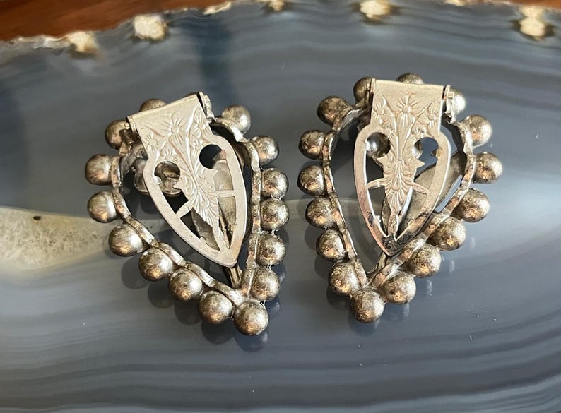 May include: A pair of silver heart-shaped earrings with a beaded border and a floral design in the center.