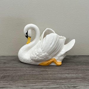May include: A white ceramic swan figurine with a curved neck and detailed feather texture. The swan has a black beak and yellow accents on its feet and beak. The figurine is a decorative item, likely for display.