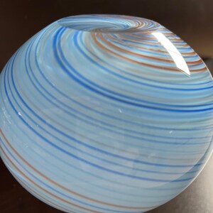 May include: A spherical glass object with a swirling pattern of blue, light blue, and orange stripes. The glass appears to be transparent, with light reflecting off the surface. The object has a rounded shape, and the stripes create a sense of movement.