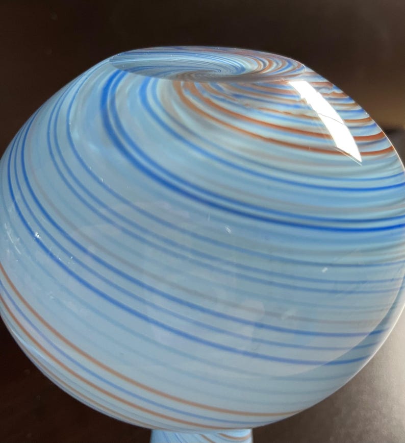 May include: A spherical glass object with a swirling pattern of blue, light blue, and orange stripes. The glass appears to be transparent, with light reflecting off the surface. The object has a rounded shape, and the stripes create a sense of movement.