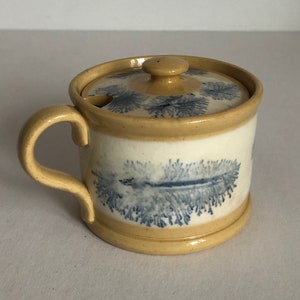 Puede incluir: Un pequeño tarro de cerámica amarillo con tapa y asa. El tarro tiene un diseño floral azul y blanco.