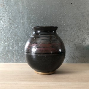 May include: A round, black ceramic vase with a brown rim and a band of pink and purple glaze around the middle.