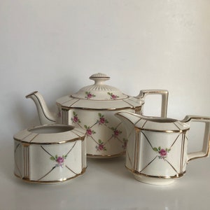 May include: A vintage tea set with a cream-coloured base and gold trim. The teapot, creamer and sugar bowl are decorated with a pink rose pattern.