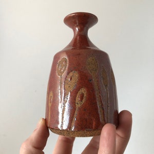 Puede incluir: Un peque&ntilde;o jarr&oacute;n de cer&aacute;mica marr&oacute;n con una superficie texturizada y un dise&ntilde;o de formas abstractas marrones y naranjas. El jarr&oacute;n tiene un cuello estrecho y una base ancha.