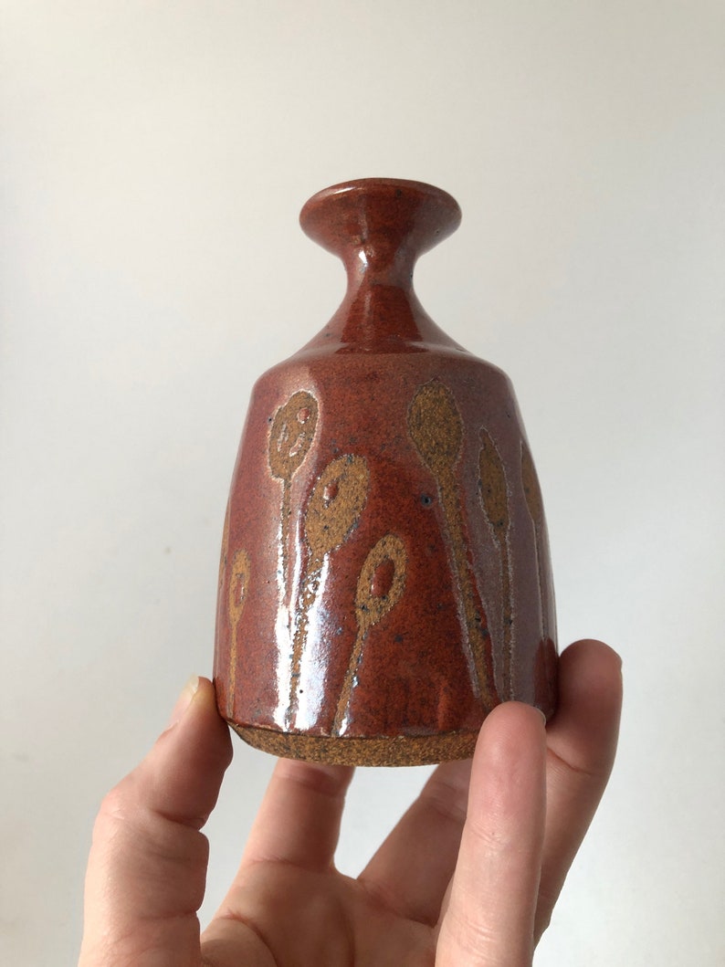 Puede incluir: Un peque&ntilde;o jarr&oacute;n de cer&aacute;mica marr&oacute;n con una superficie texturizada y un dise&ntilde;o de formas abstractas marrones y naranjas. El jarr&oacute;n tiene un cuello estrecho y una base ancha.