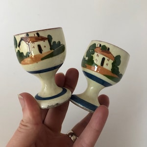 Op de afbeelding: Twee witte keramische eierdopjes met een blauw en bruin geverfd ontwerp van een huis en bomen.