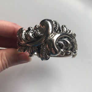 K&ouml;nnte beinhalten: Ein silbernes Armband mit einem detaillierten, verzierten Design mit wirbelnden, blattartigen Mustern.