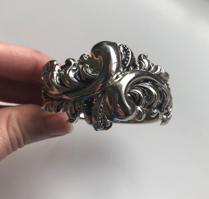 K&ouml;nnte beinhalten: Ein silbernes Armband mit einem detaillierten, verzierten Design mit wirbelnden, blattartigen Mustern.