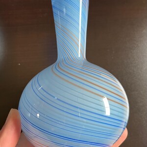 May include: A hand-blown glass vase with a rounded base and a slender neck. The vase features a light blue background with swirling stripes of darker blue and orange. The vase is approximately 8 inches tall.
