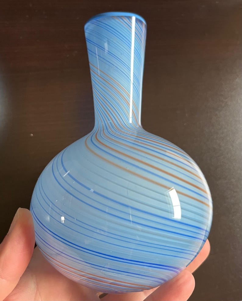 May include: A hand-blown glass vase with a rounded base and a slender neck. The vase features a light blue background with swirling stripes of darker blue and orange. The vase is approximately 8 inches tall.