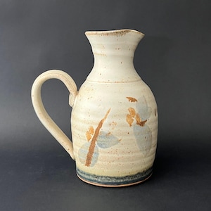 May include: A handmade ceramic pitcher with a cream-colored base and a curved handle. The pitcher features brown and blue floral designs and a dark blue band near the base. The pitcher is approximately 8 inches tall.