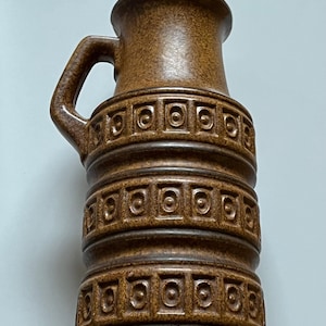 May include: A brown ceramic vase with a handle. The vase has a textured surface and is decorated with repeating square patterns. The vase is approximately 10 inches tall.
