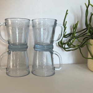 Peut inclure: Trois tasses en verre transparent avec une teinte bleu clair et un motif gravé décoratif. Les tasses ont des poignées et sont empilées les unes sur les autres.