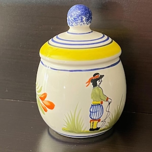 May include: A ceramic jar with a lid, featuring a hand-painted design. The lid has blue and yellow stripes and a blue knob. The jar's body is white with a painted scene of a person in traditional attire and orange flowers.